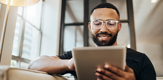 Man wearing white rimmed glasses sitting on a sofa looking at a tablet